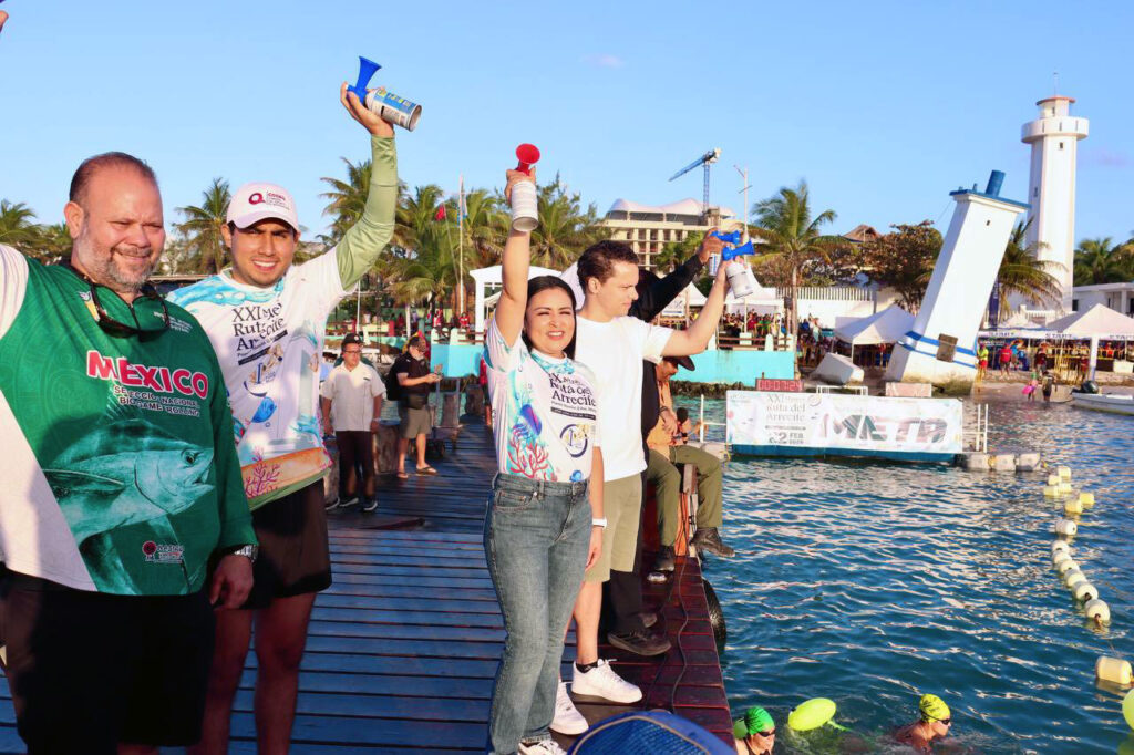 Race officials stand on the Puerto Morelos pier holding air horns above the water as swimmers gather near the start line, with the lighthouse behind.