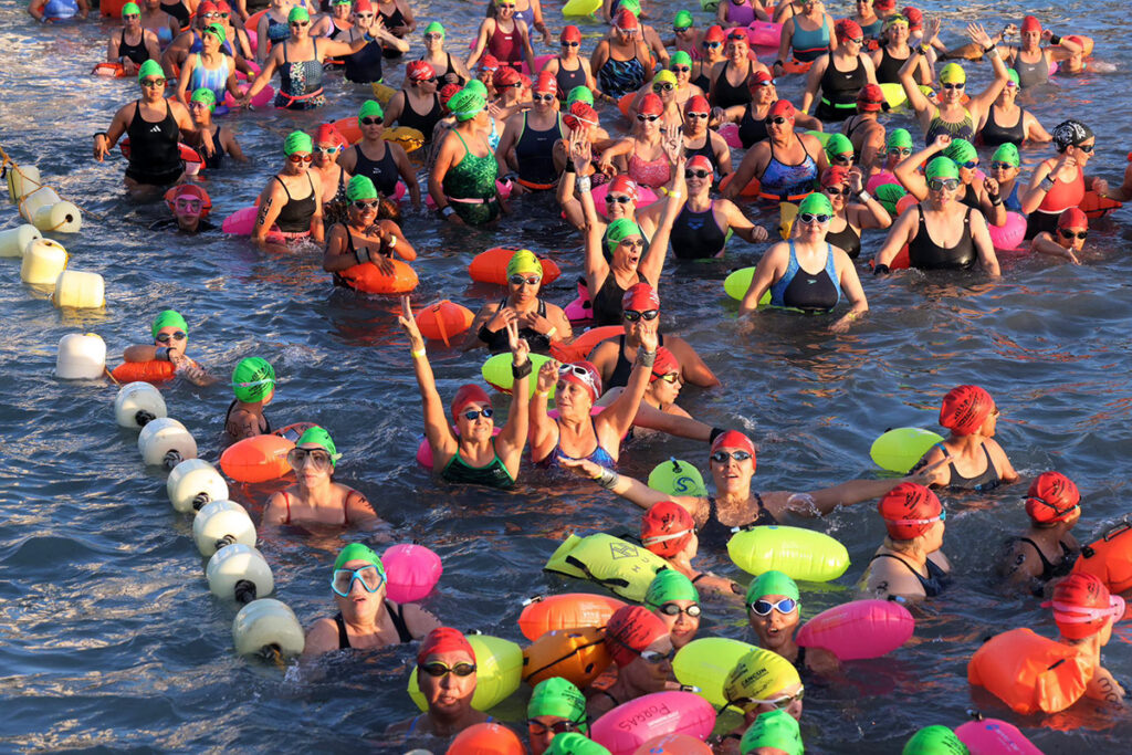 Crowd of open-water swimmers in colorful caps and tow-floats waits in the water beside a line of white buoys at the start.