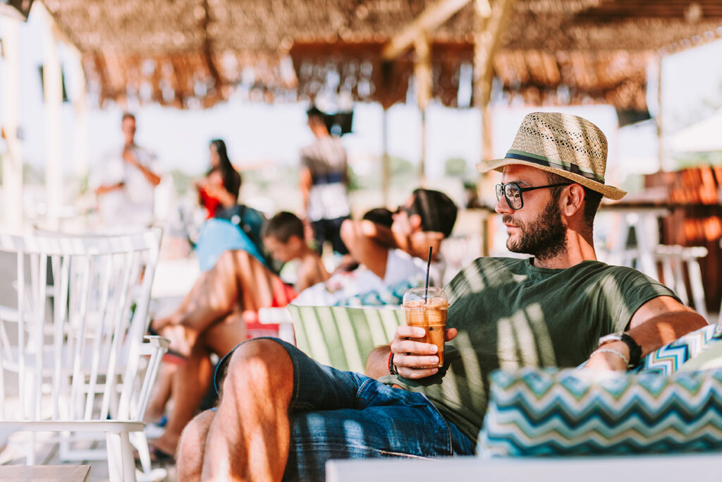 Man in a hat relaxes on a striped cushion with a drink in hand at a beach club, with other guests and palapa shade behind.