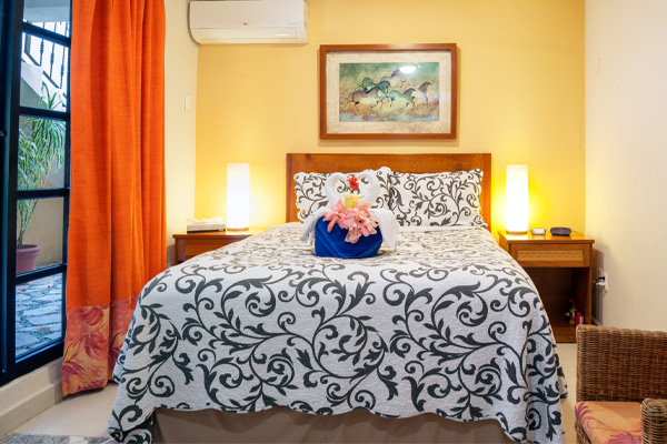 Cozy bedroom with patterned bedspread, warm lamps, orange curtains, and an AC unit above the bed.