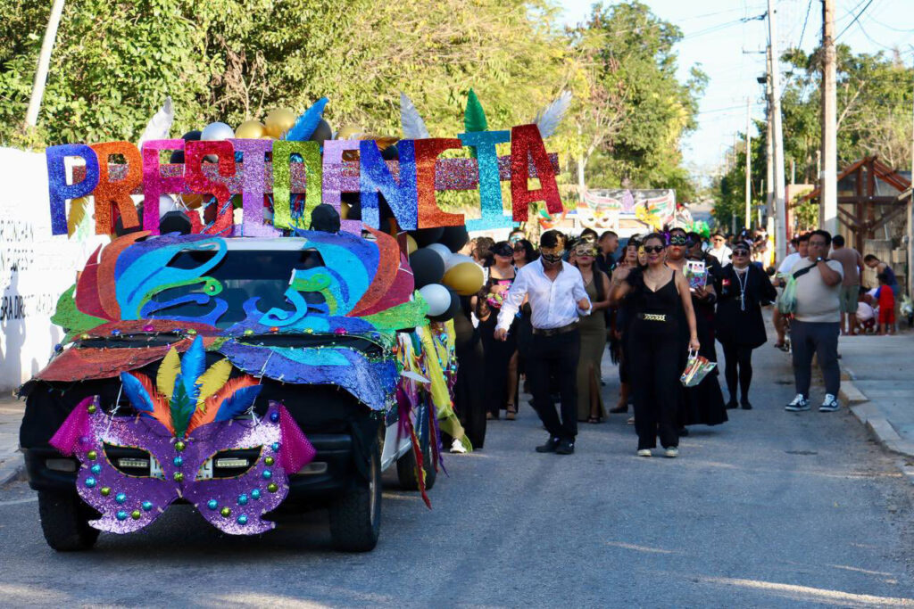 A colorful Carnival float with a large “PRESIDENTA” sign leads a parade down a sunlit street.