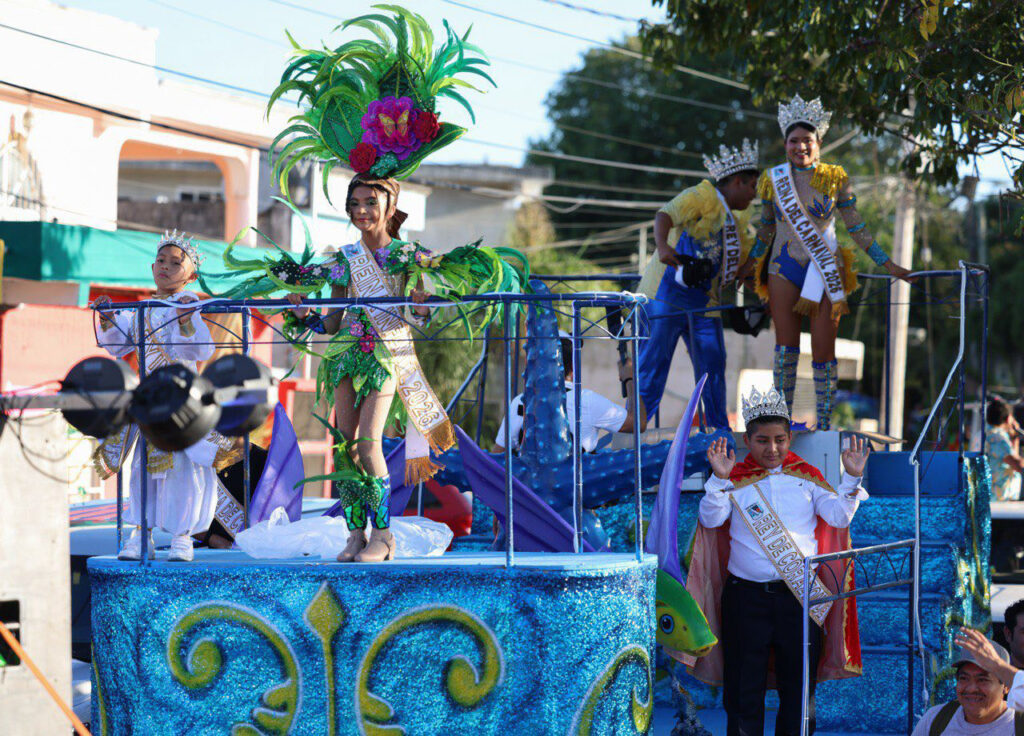 Crowned Carnival court in bright costumes stands on a glittery blue float while a child waves from the front.