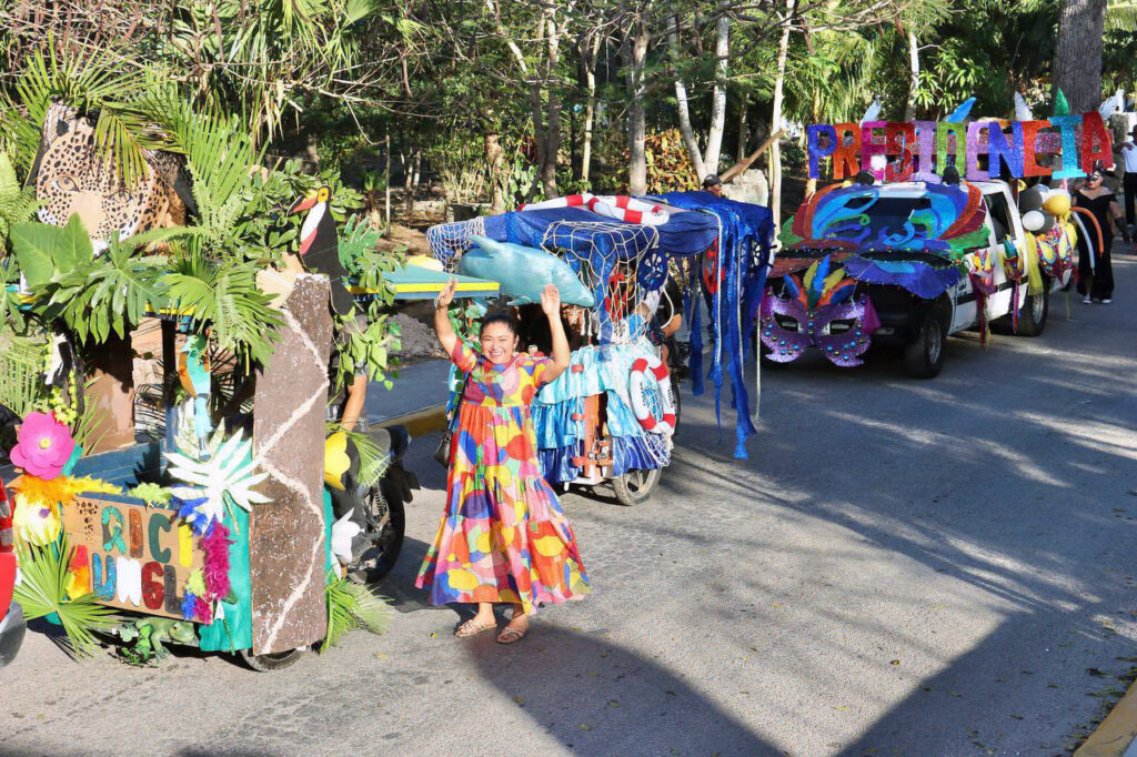 A woman in a colorful dress waves beside a jungle-themed float as more Carnival floats line up behind her.