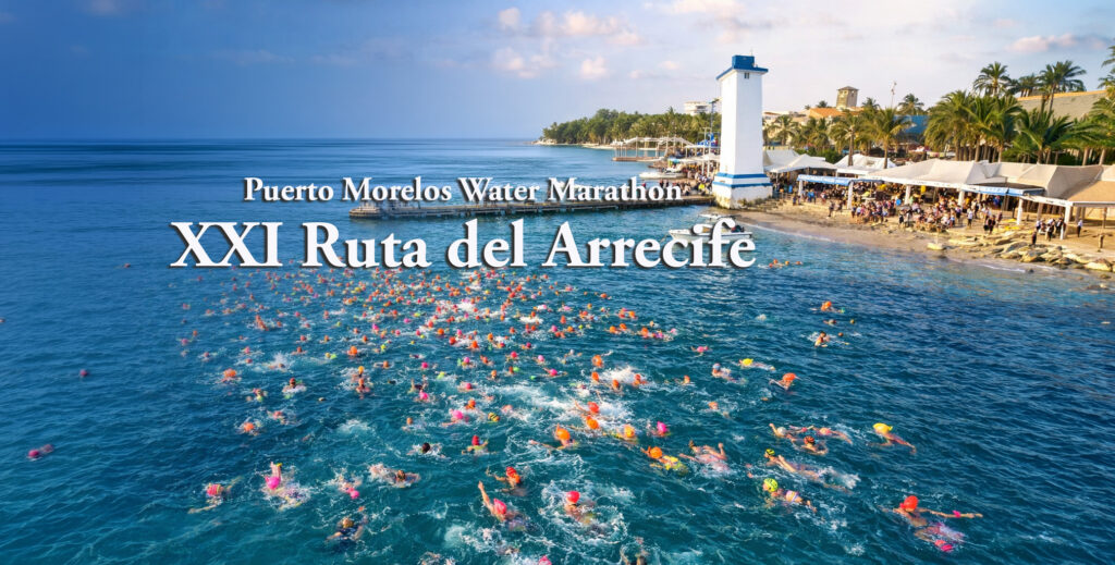 Aerial view of swimmers in colorful caps crossing turquoise water near the Puerto Morelos pier and lighthouse during Ruta del Arrecife.