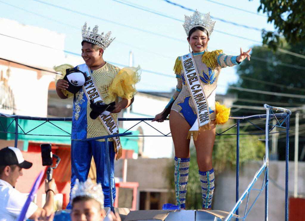 Carnival king and queen in crowns and sashes wave from a parade float.