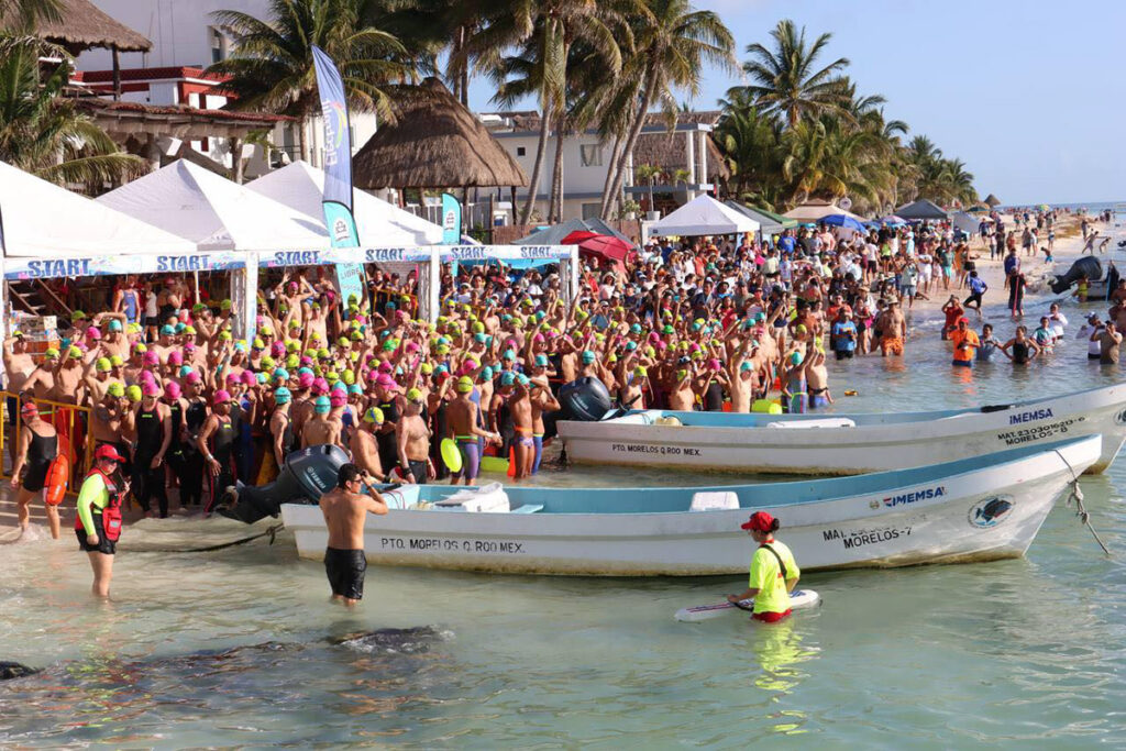 Large crowd of swimmers and spectators gathers on the Puerto Morelos beach at the start line, with small boats anchored in the shallows.