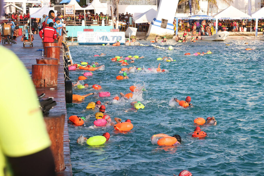 Swimmers in colorful caps and tow-floats crowd the water near the pier as safety staff watch along the edge at the Ruta del Arrecife event.