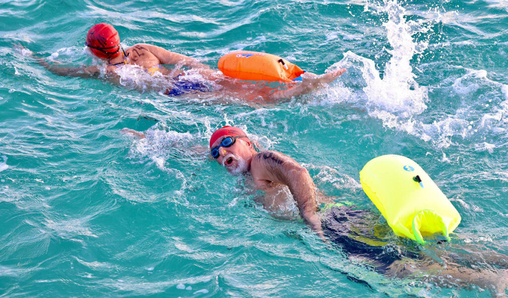 Two open-water swimmers move through turquoise water with safety tow-floats, one looking toward the camera mid-stroke.