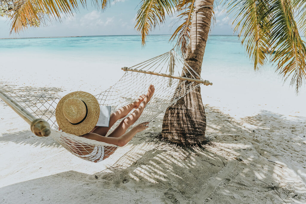 A person relaxes in a hammock under palm trees on a quiet white-sand beach with turquoise water nearby.