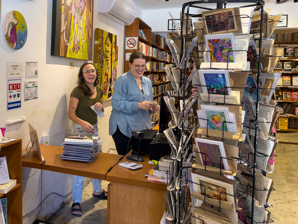 Two staff members behind the counter at Alma Libre Books & Gifts, surrounded by shelves and racks of books and postcards.