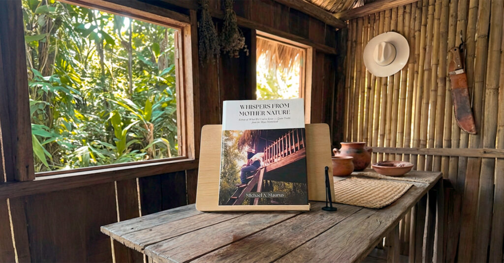 A copy of Whispers from Mother Nature stands on a rustic table beside a mug, framed by an open window looking into the jungle.