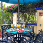 Shaded patio table set for breakfast with fruit and juice, surrounded by tropical plants and an umbrella.