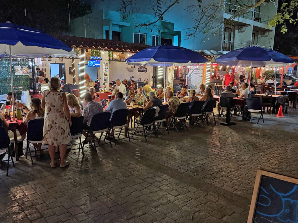People gather at outdoor tables and restaurants in Puerto Morelos on a lively evening by the town square.