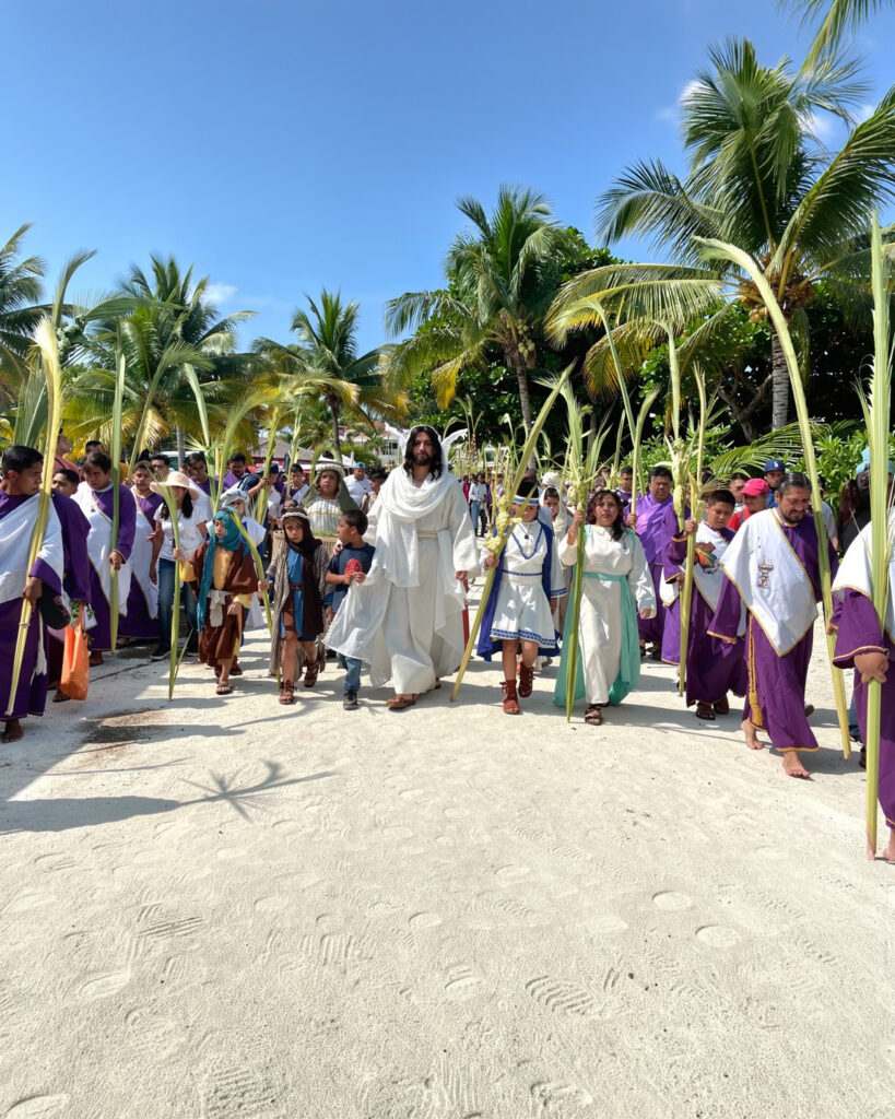 People in biblical costumes walking in a Holy Week procession lined with palm leaves outdoors.