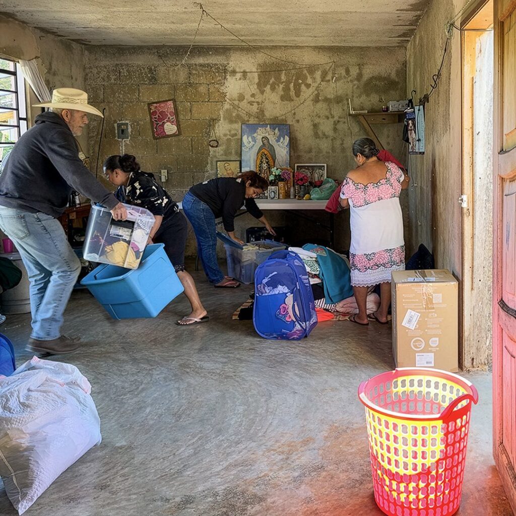 Michael A. Maurus brings donated items into a modest home in a Maya village while a woman and child stand nearby.