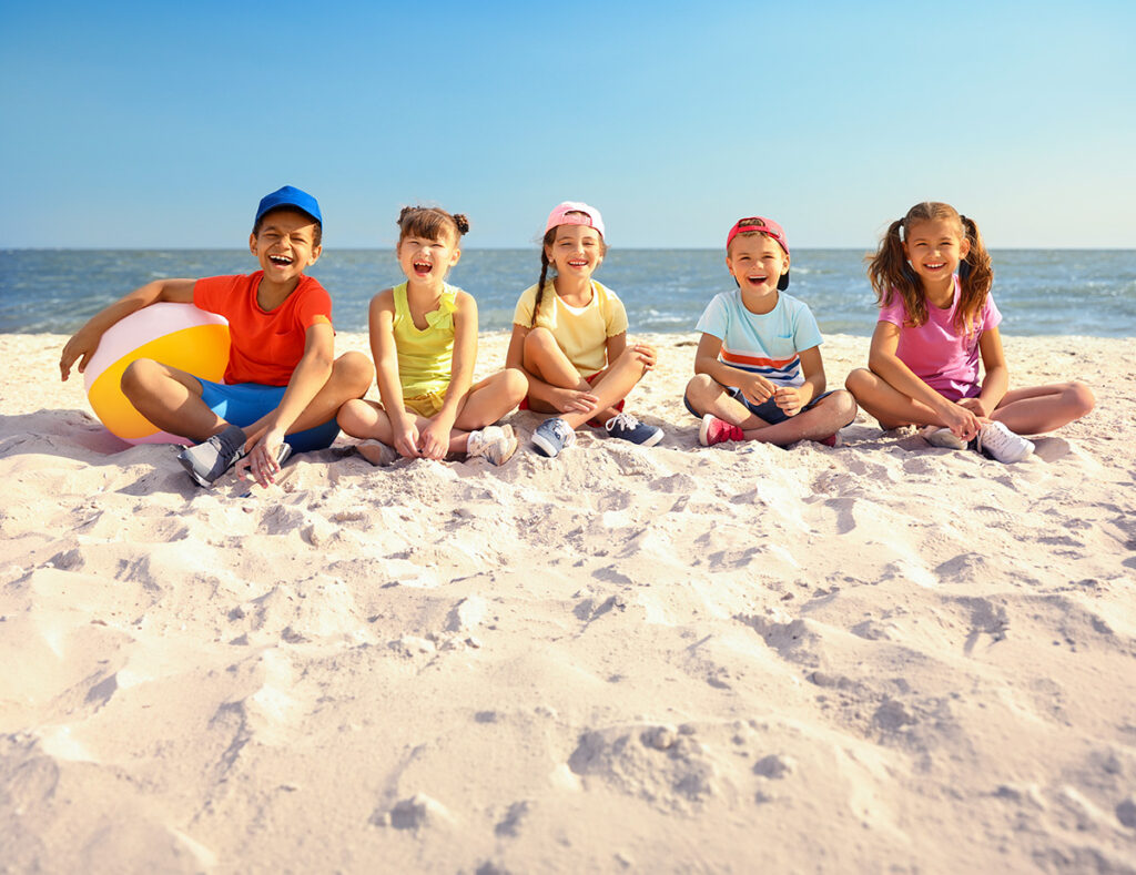 Five children sitting together on a sunny beach, smiling toward the camera with the sea behind them