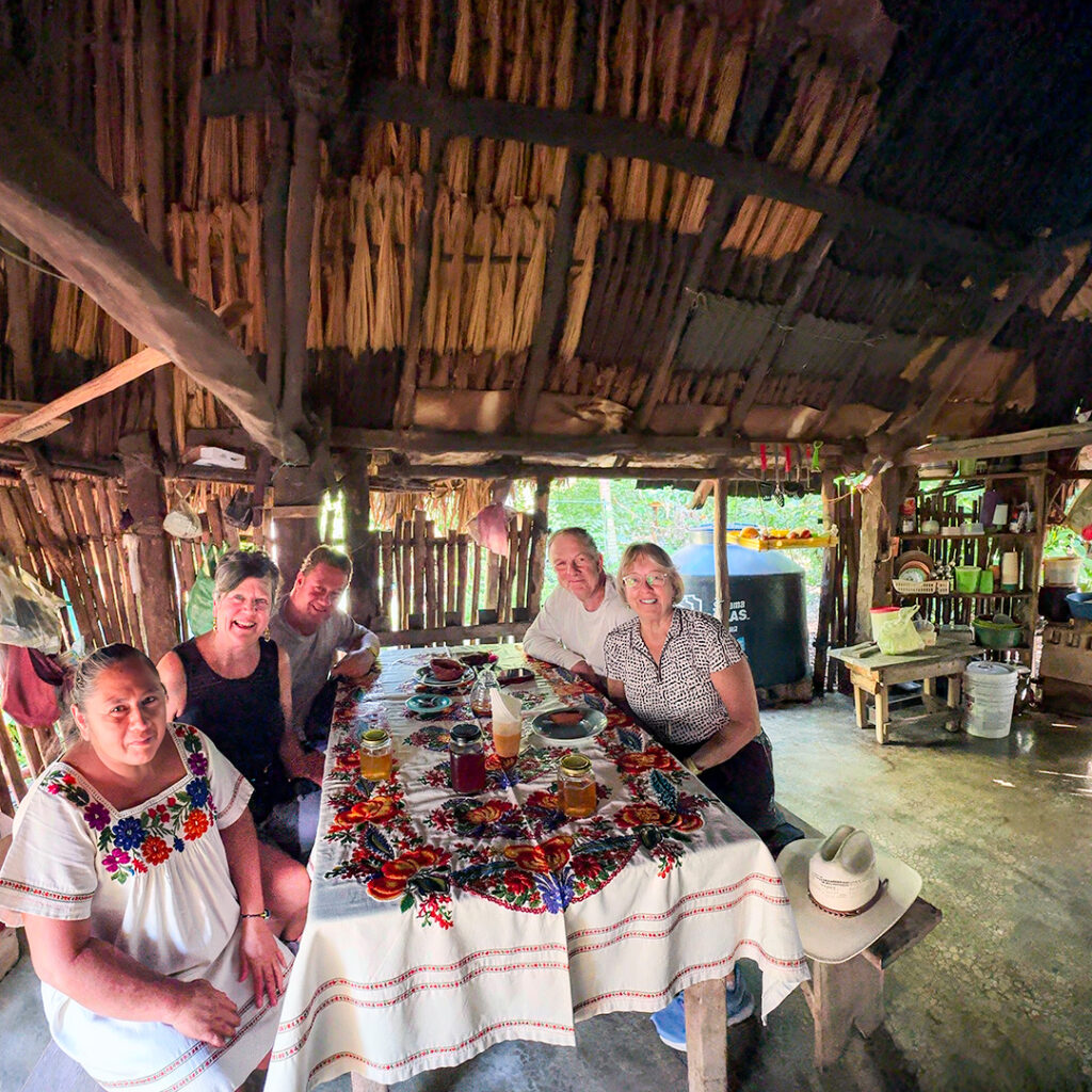 Guests and Maya artisans gather around a table of handmade crafts inside a traditional palapa.