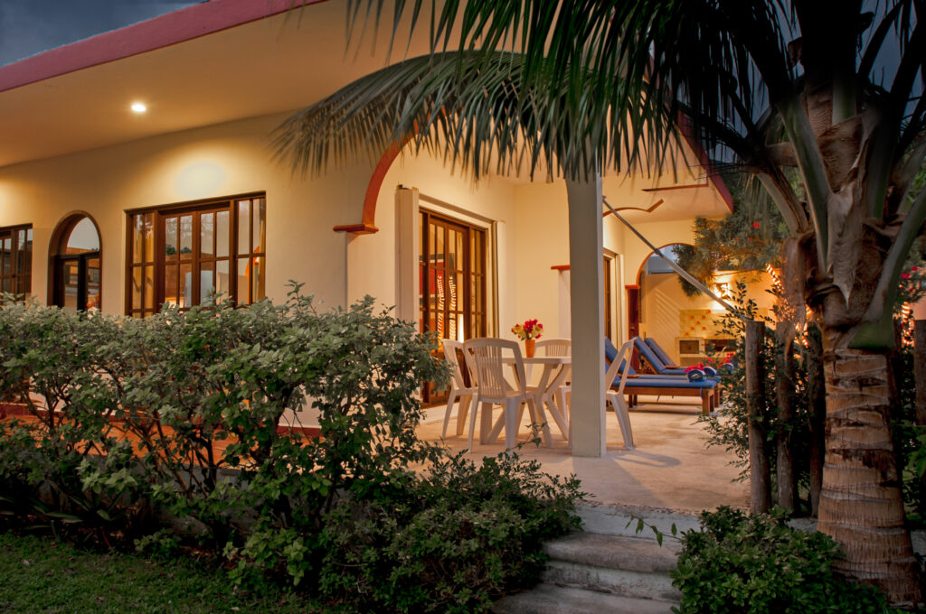 Garden shrubs and a palm beside a lit patio with outdoor chairs and lounge seating.