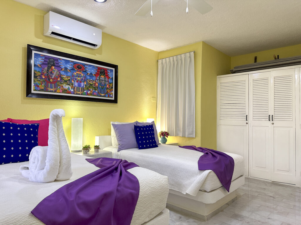 Beds with purple throws beside white closet doors beneath framed colorful artwork.