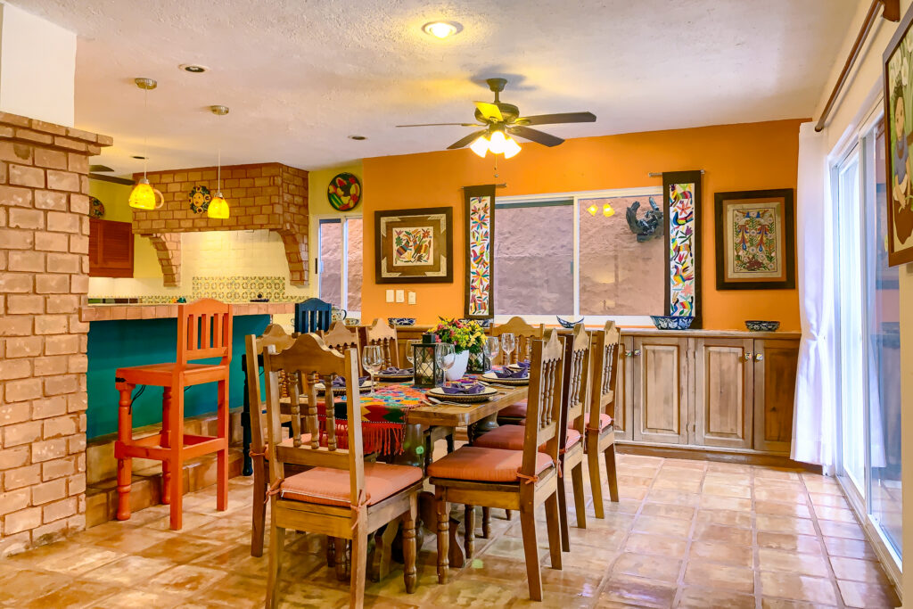 Long dining table with wooden chairs beside a tiled kitchen bar and orange accent walls.