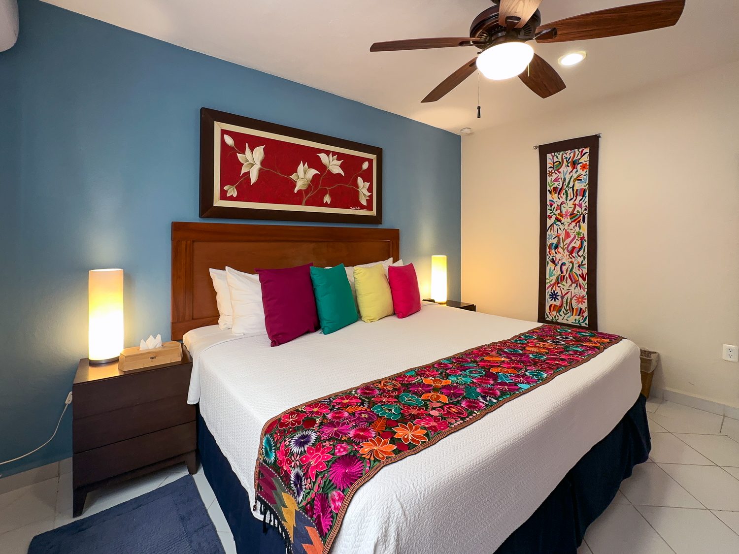 White bed with bright pillows and a floral runner beneath red artwork on a blue wall.