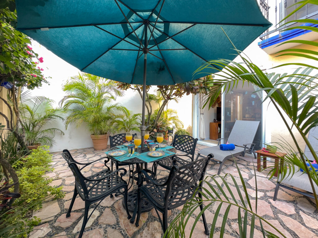 Outdoor dining set and reclining chairs on a stone patio enclosed by tropical plants.