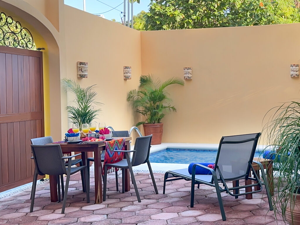 Outdoor dining table and lounge chair beside the pool against yellow walls with potted palms.