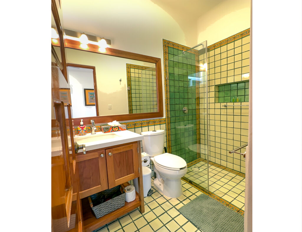 Wooden vanity, mirror, toilet, and glass shower surrounded by yellow-and-green tiles.