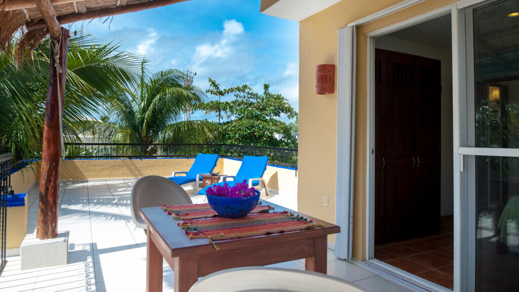 Wooden terrace table beneath thatch with blue lounge chairs beyond among palms.