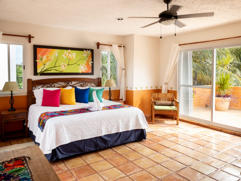 Bed with colorful pillows and a floral runner beneath green artwork beside wide terrace doors.