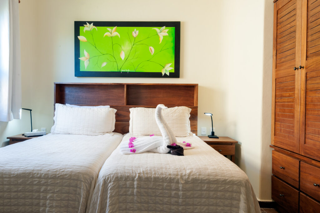 Two beds with white covers beneath green artwork beside a tall wooden wardrobe.