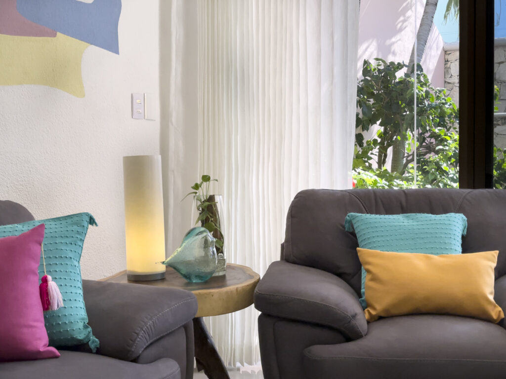 Gray armchairs with turquoise and yellow cushions beside a small table and curtained windows.