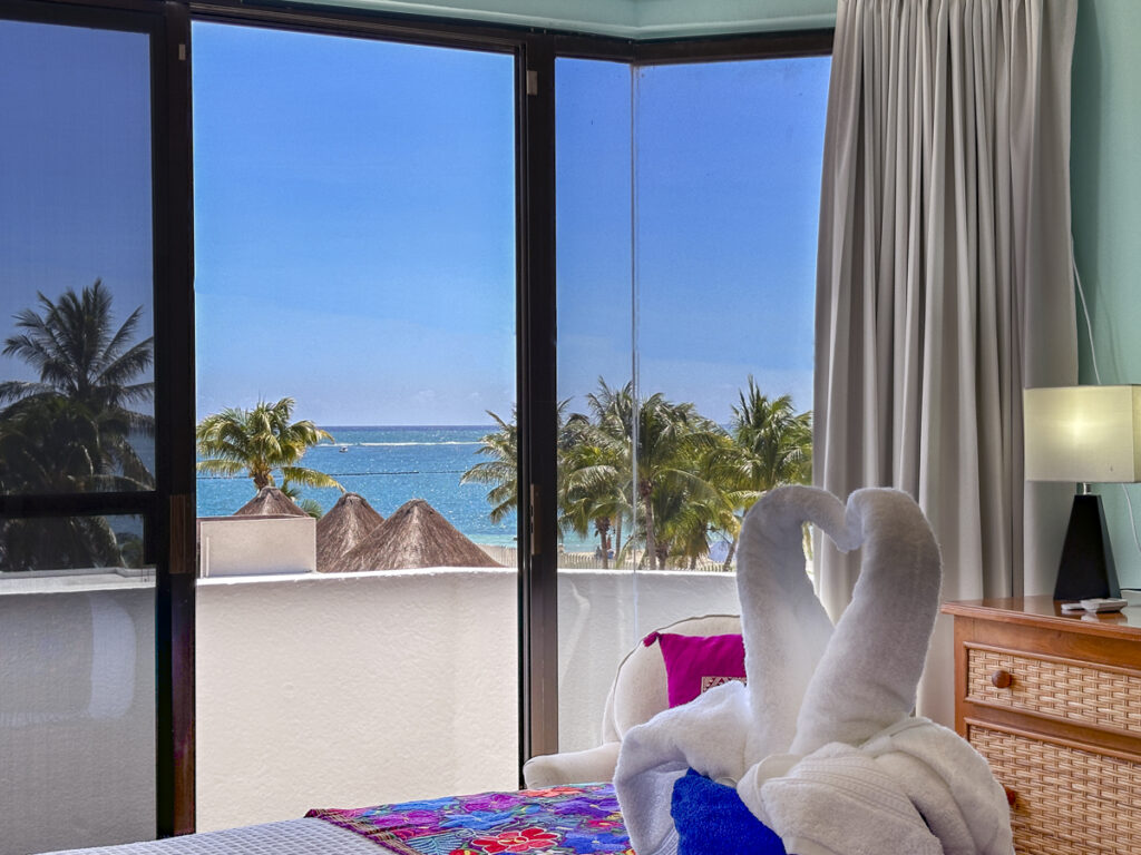 Towel swans on the bed in front of windows overlooking palms and turquoise sea.