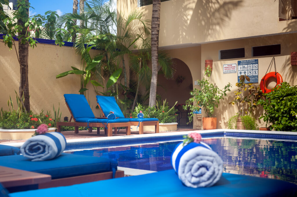Rolled towels on blue lounge chairs beside the pool, with yellow courtyard walls and palms beyond.