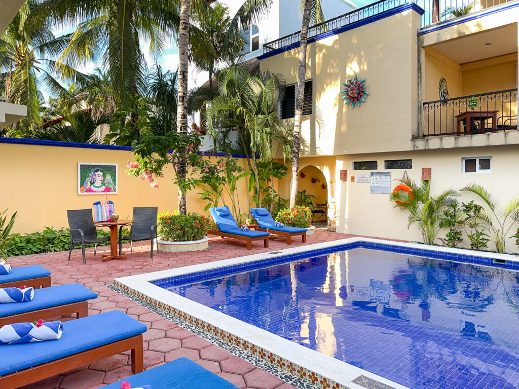 Blue swimming pool and lounge chairs beneath palms beside a yellow building with balconies.