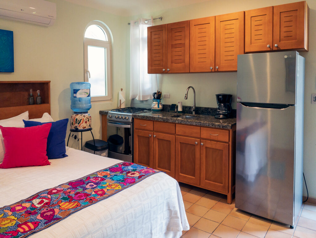 Bed with colorful bedding beside a kitchen with wooden cabinets, stove, sink, and refrigerator.
