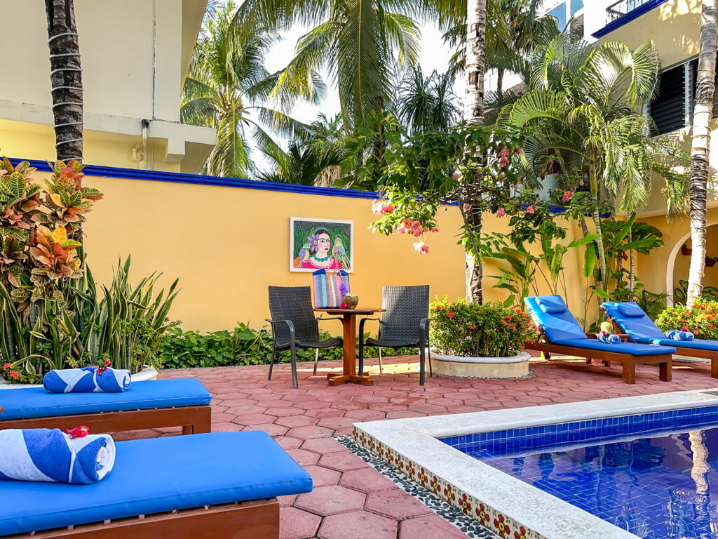 Blue lounge chairs and a small table beside the pool, with palms and artwork on a yellow courtyard wall.