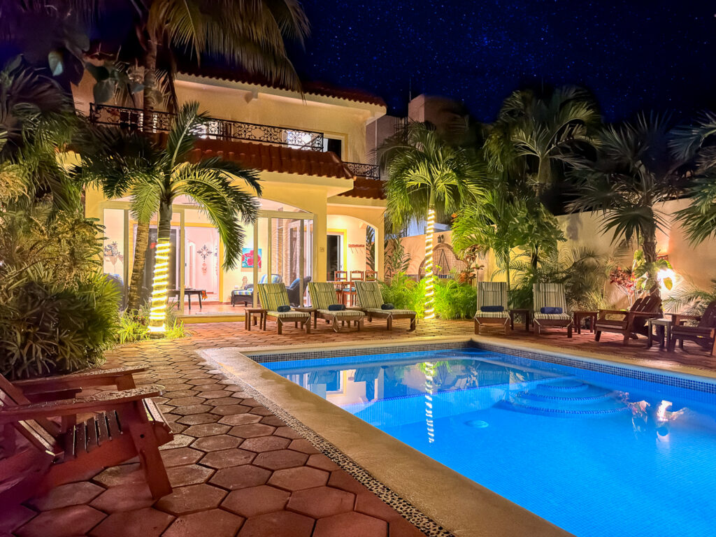 Illuminated blue pool in front of a yellow house surrounded by palms at night.