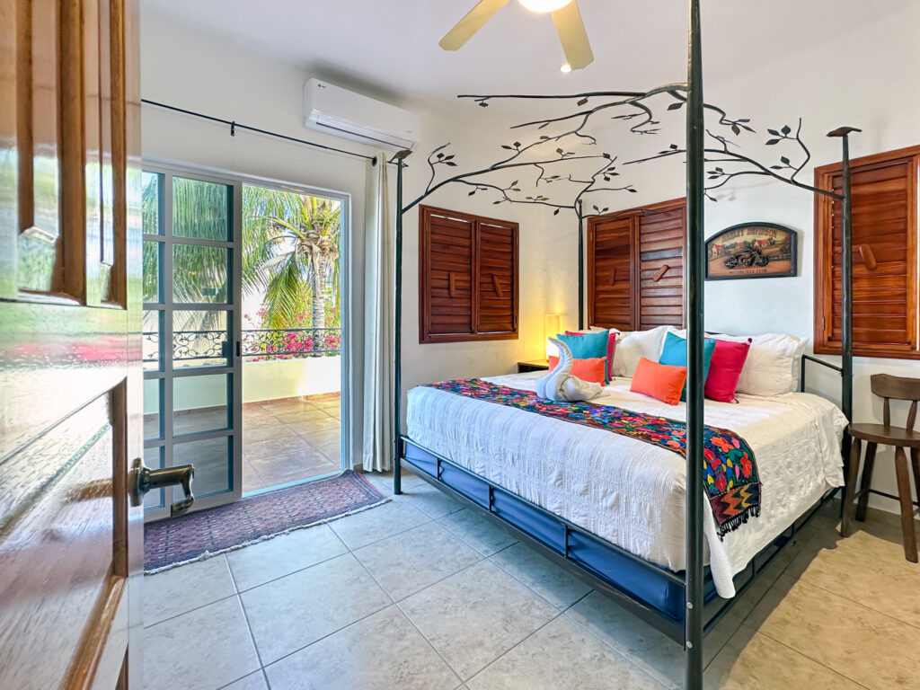 Four-poster bed with colorful pillows and a floral runner beside balcony doors and painted branches on the wall.