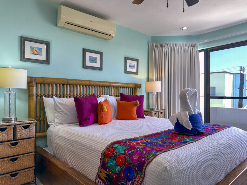 Bed with orange and purple pillows and a floral runner in a turquoise bedroom near wide windows.