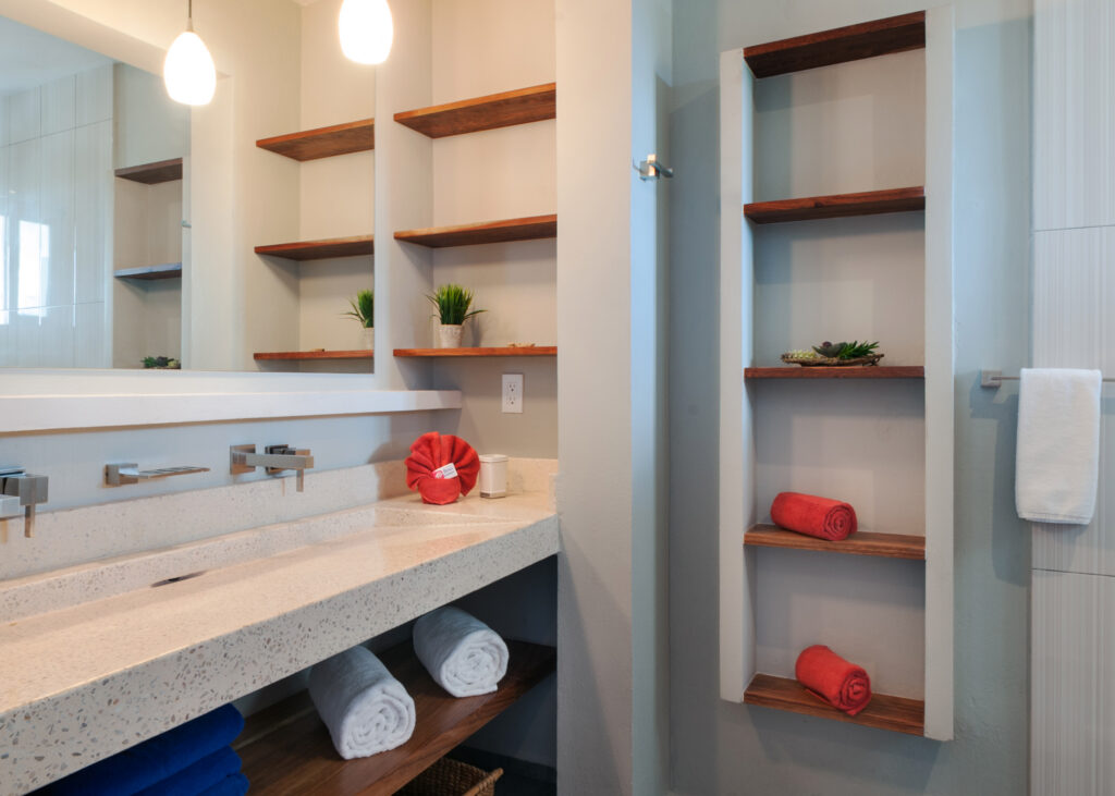 Long bathroom counter beneath a wide mirror beside open shelves holding towels and small plants.