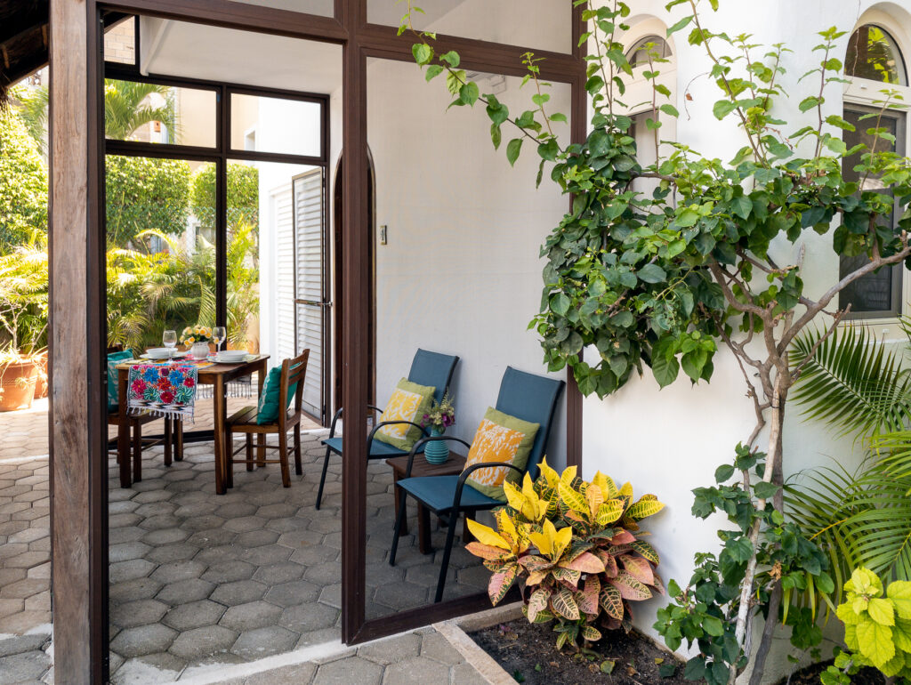 Open screened door reveals a patio table and blue chairs, surrounded by leafy plants.