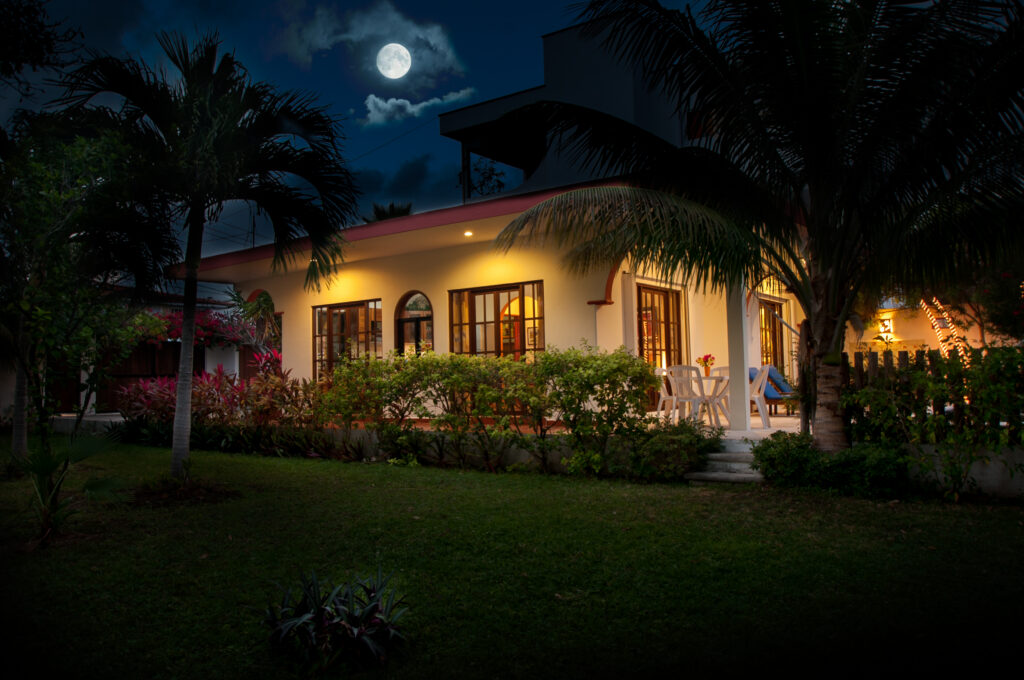 Illuminated low-rise house beneath palms and a moonlit sky, surrounded by lawn and shrubs.