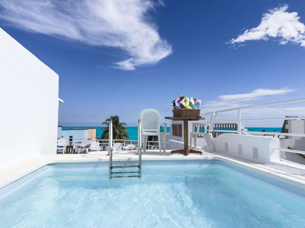 Small rooftop pool beside a dining table beneath blue skies with the sea in the distance.
