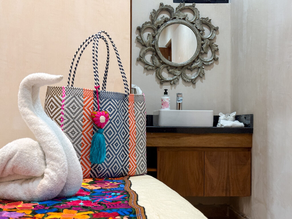 Towel swan and patterned woven bag on a floral bed runner near a bathroom vanity.