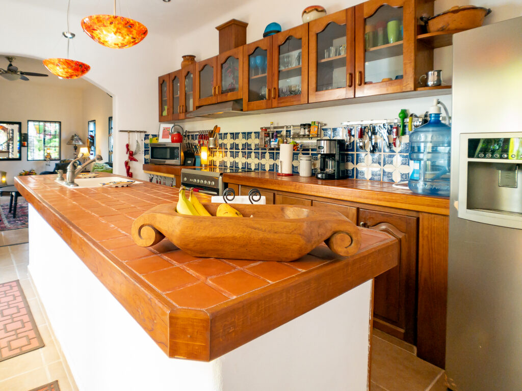 Tiled kitchen island with a wooden bowl beside cabinets, open shelves, and stainless steel refrigerator.