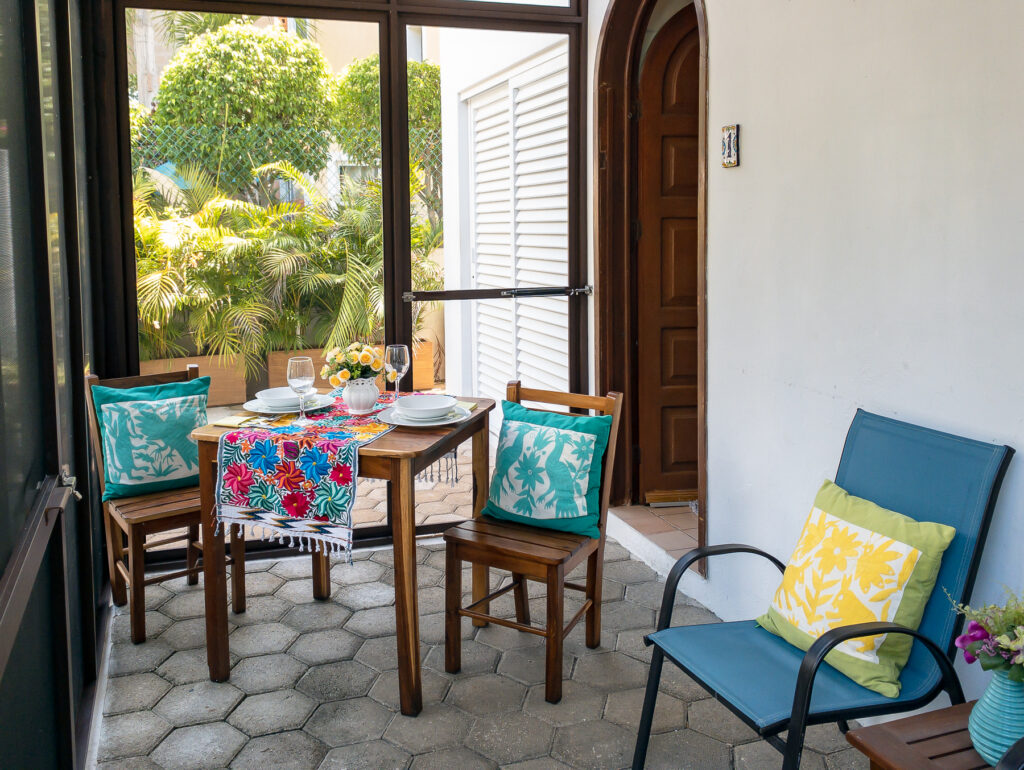 Table set for two beside blue chairs in a screened patio with garden views.