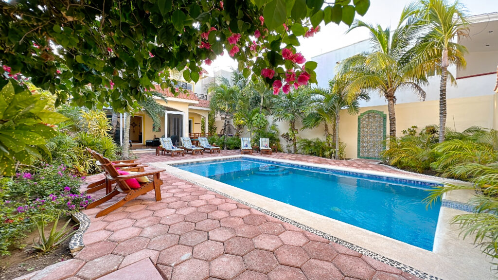 Long rectangular pool surrounded by red paving, lounge chairs, flowering plants, and palms.