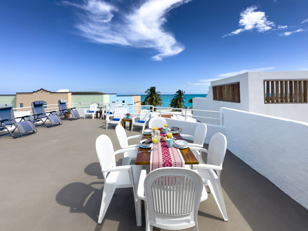 Long table set for dining on an open rooftop terrace with lounge chairs and sea views.