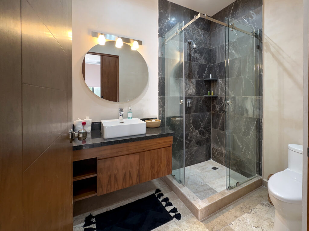 Wooden vanity with rectangular basin and round mirror beside a glass-enclosed shower with dark tiles.